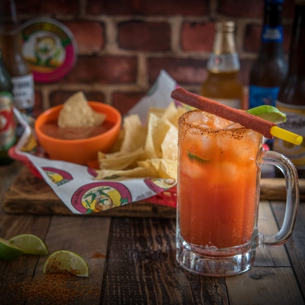 A Guanataco michelada with a tamarind candy straw, chips, and salsa on a wooden table.