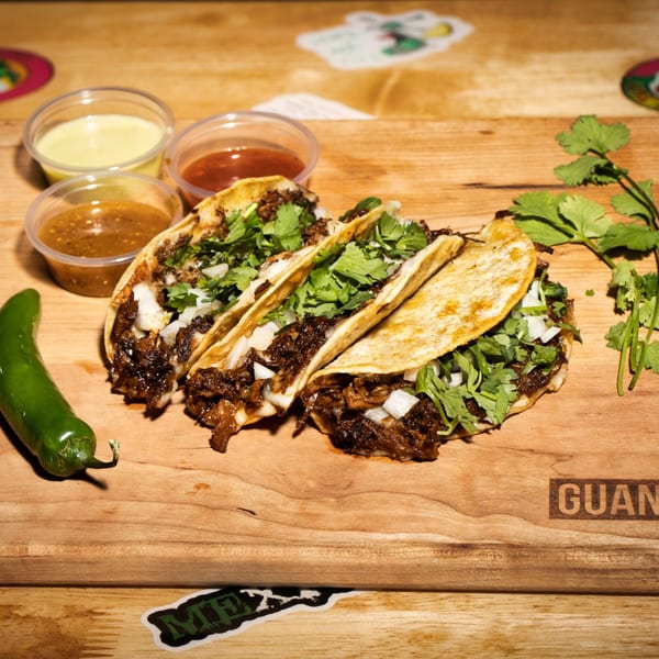 Three Guanataco tacos with meat, cilantro, and onions, served with sauces and a jalapeño.