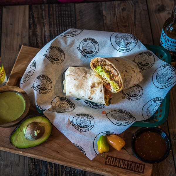 Two Guanataco burritos, one cut open, with avocado, sauces, and a Corona beer on a wooden board.