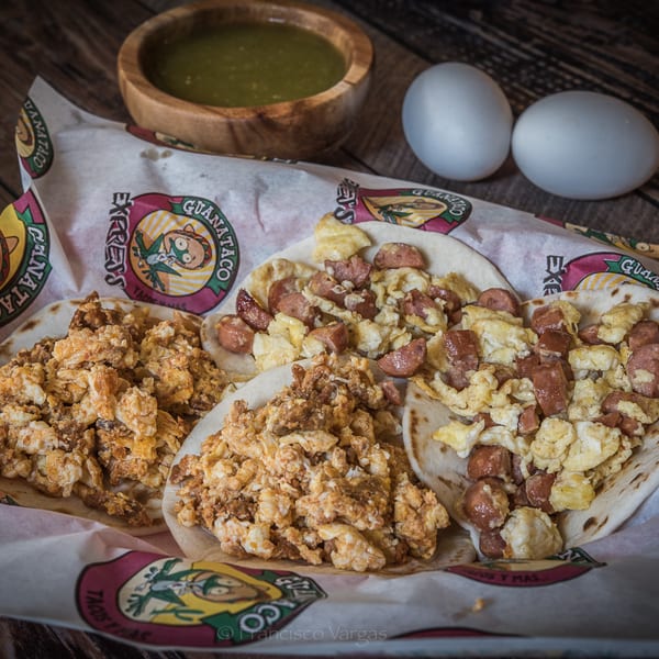 Guanataco breakfast tacos with scrambled eggs and sausage, salsa, and two eggs on a wooden table.