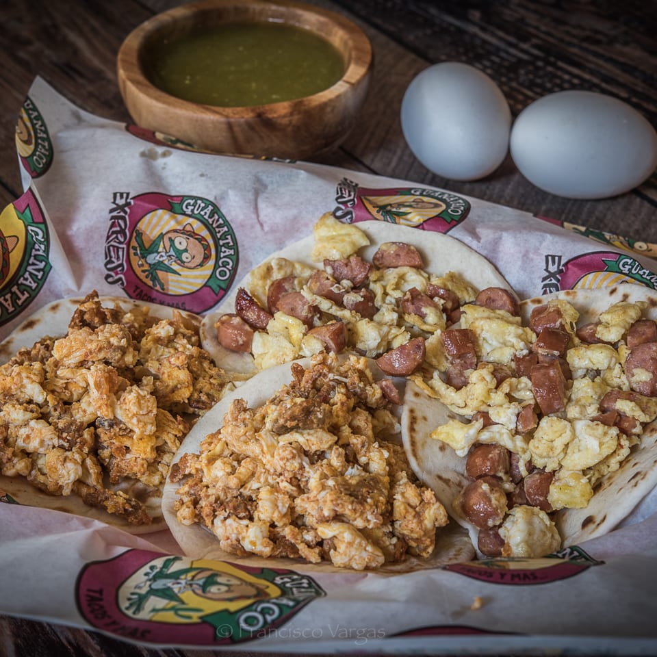 Guanataco breakfast tacos with scrambled eggs and sausage, salsa, and two eggs on a wooden table.