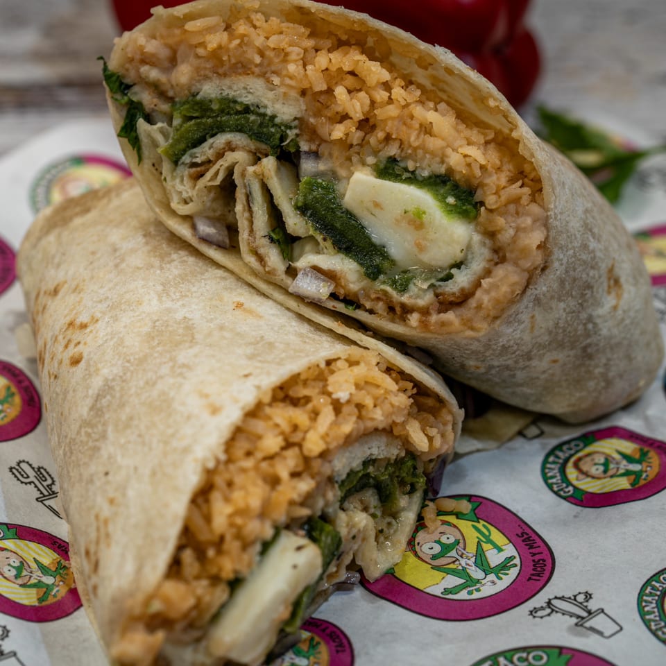 Two Guanataco burritos, one cut in half, revealing rice, green chiles, and cheese on branded paper.