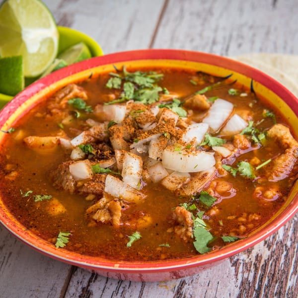 Close-up of Guanataco's menudo soup with cilantro, onions, and spices, served with lime wedges.